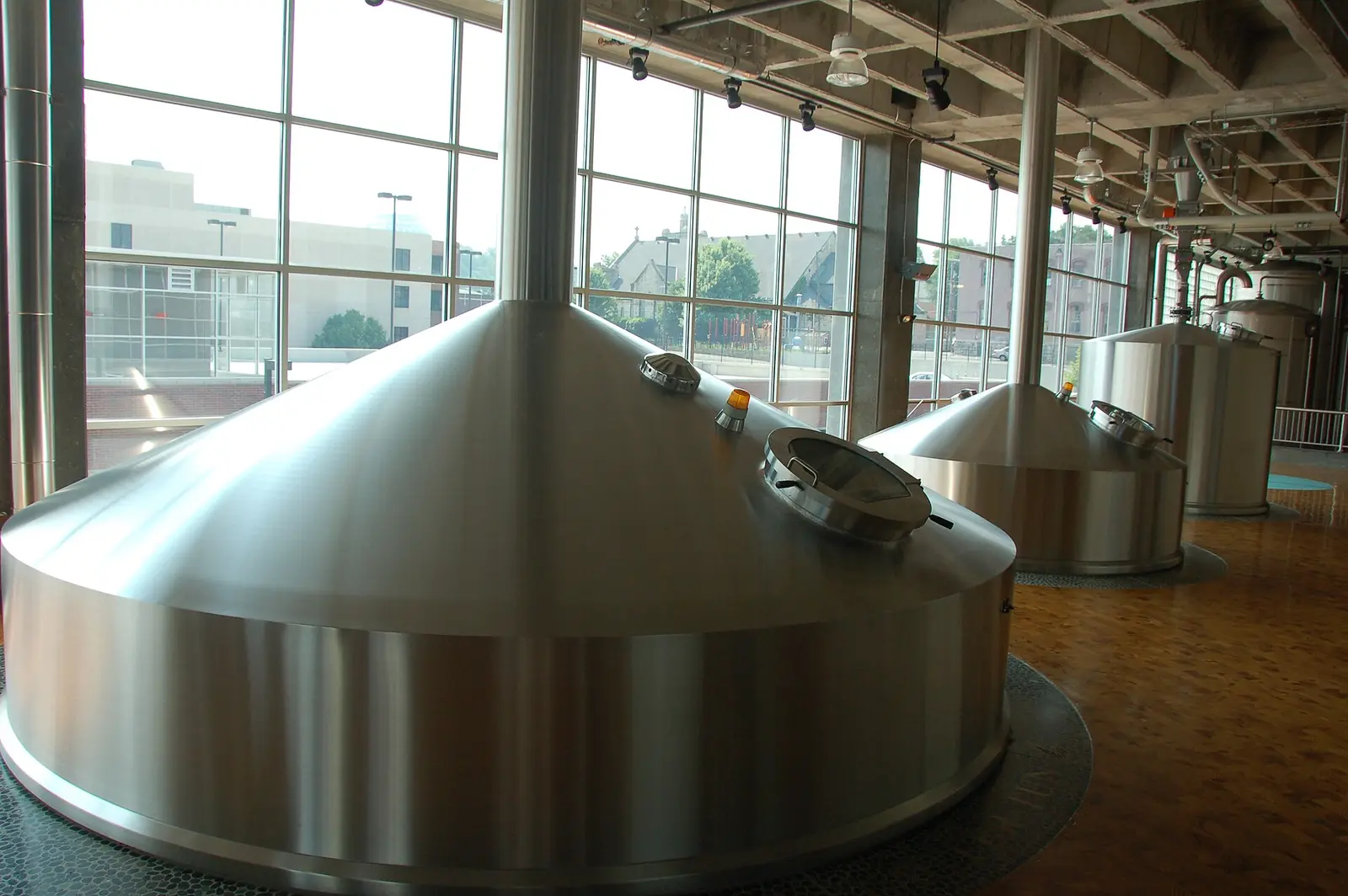 Brewhouse with three large stainless-steel brew vessels with conical tops and manway hatches, standing on a tiled floor beside tall windows