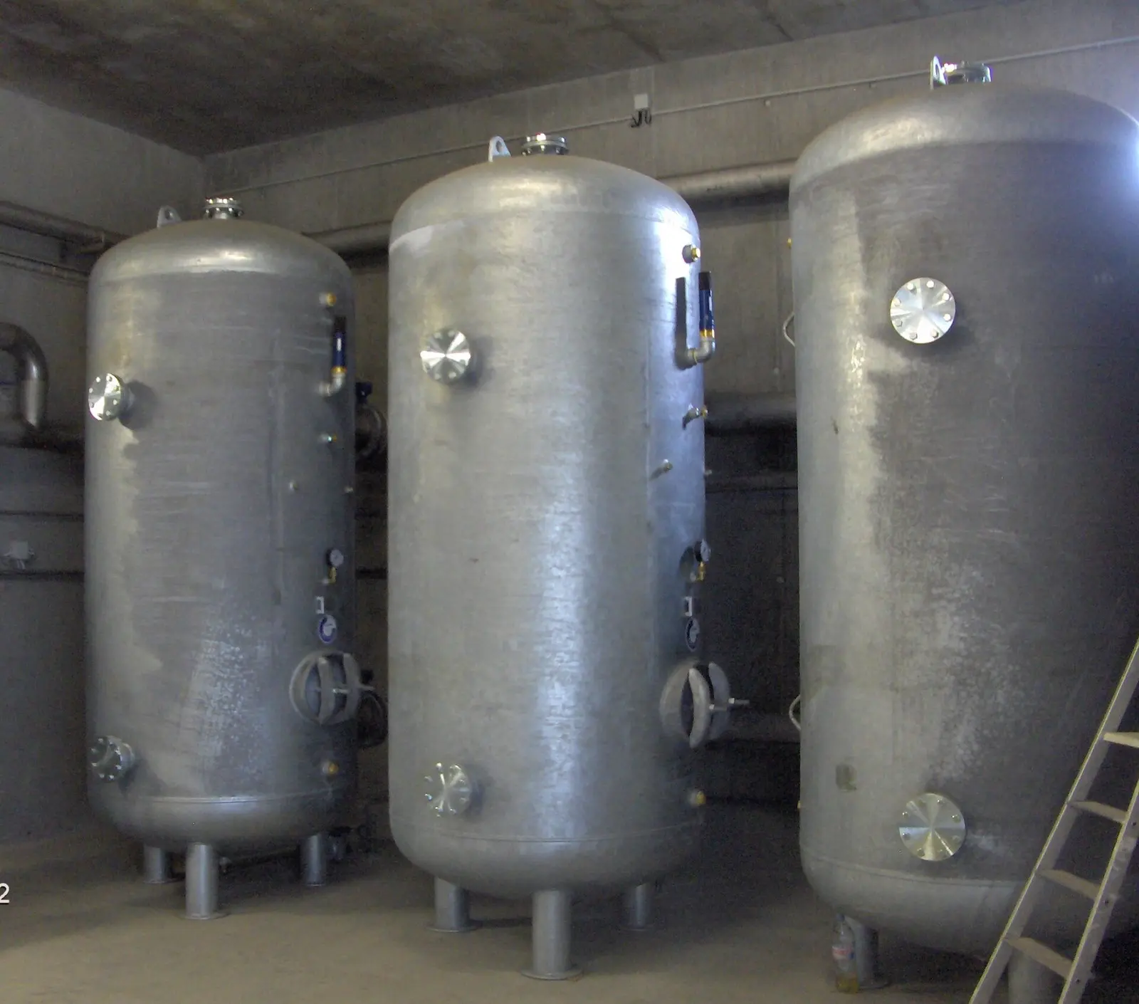 Three large vertical galvanised steel compressed air receiver tanks on legs in a concrete plant room, fitted with inspection covers and gauges