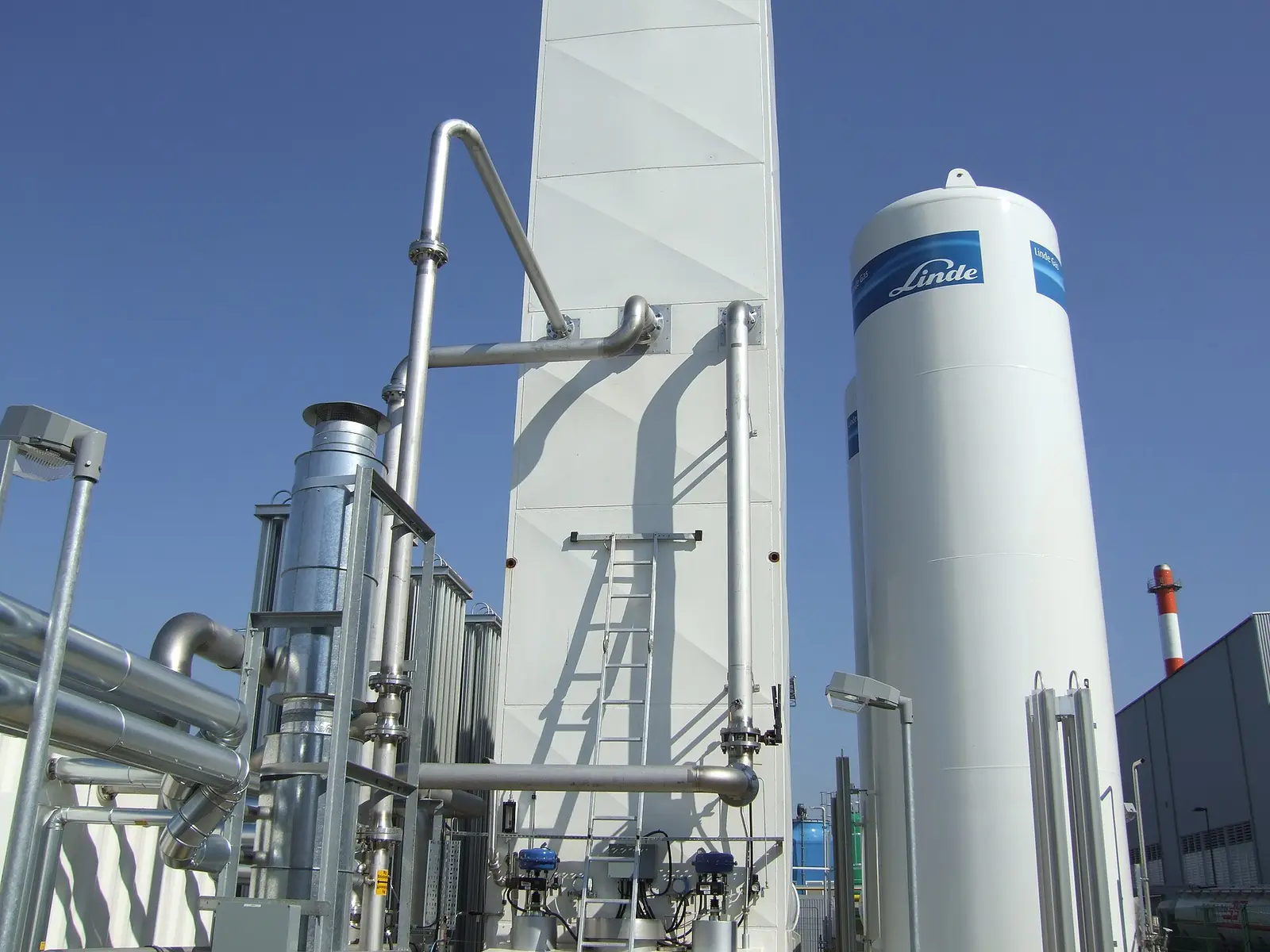 Tall white cold box of an air separation plant with stainless-steel process piping and a ladder, beside a vertical liquid gas storage tank, against a blue sky