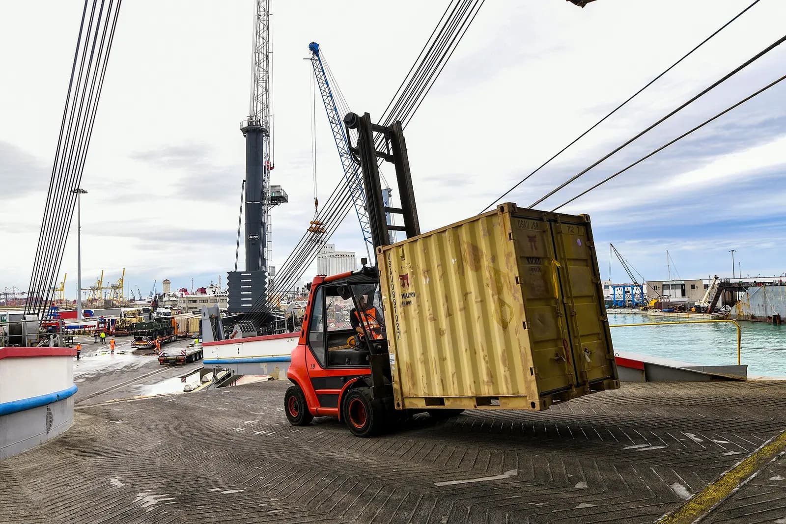 Red counterbalance forklift truck lifting a small tan shipping container on a port quayside, with cranes and the harbour in the background