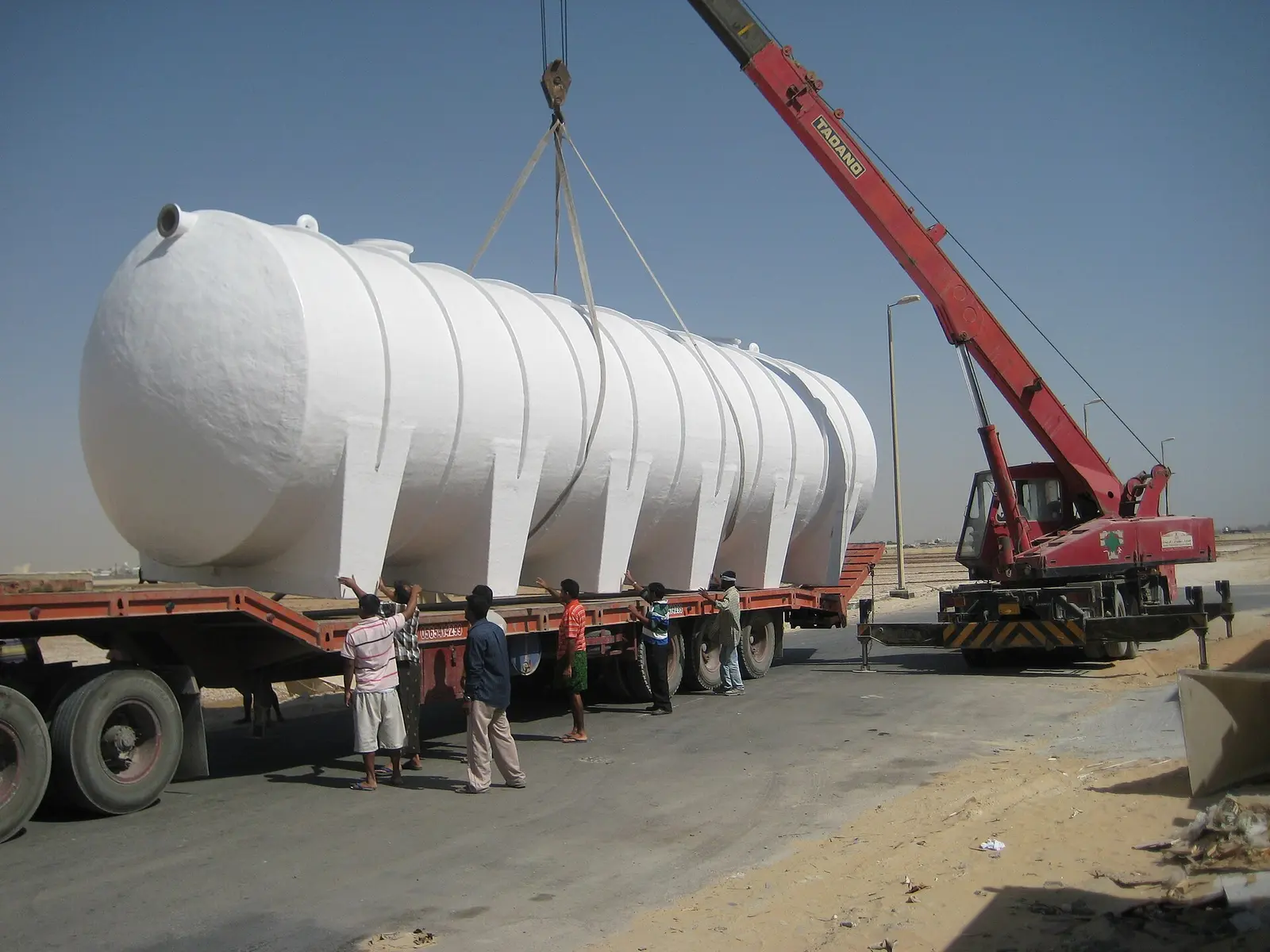 Large horizontal cylindrical fibreglass-clad storage tank, shown on a low-loader trailer being lifted by a mobile crane