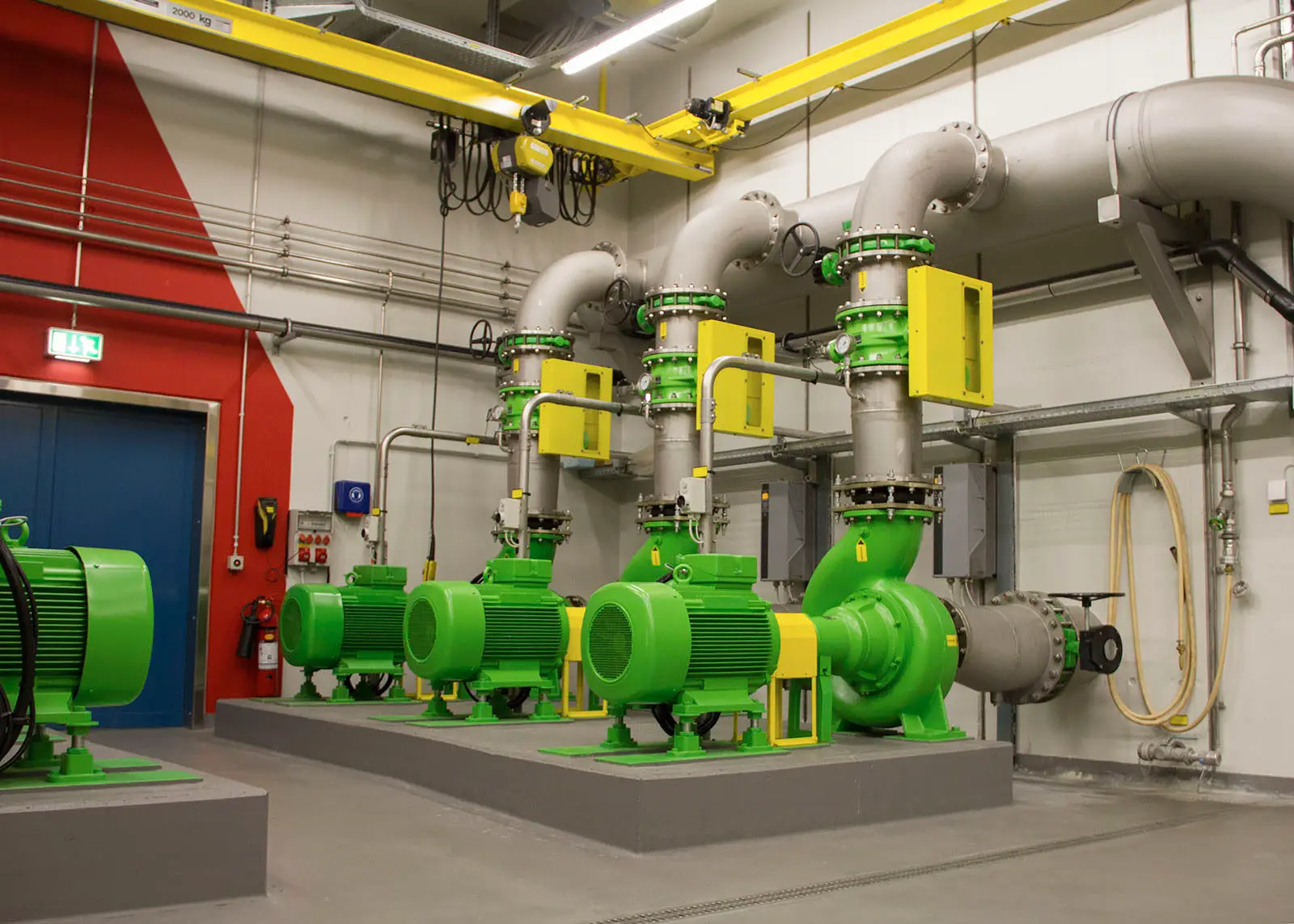 Row of green horizontal end-suction centrifugal pumps with electric motors mounted on baseplates on a concrete plinth, connected to vertical stainless-steel pipework in a pump room