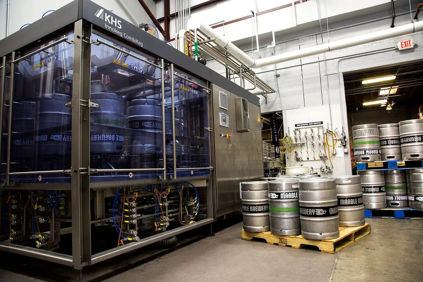 Keg washer and filler, an enclosed stainless keg cleaning and filling machine with kegs inside and filled kegs on a pallet beside it