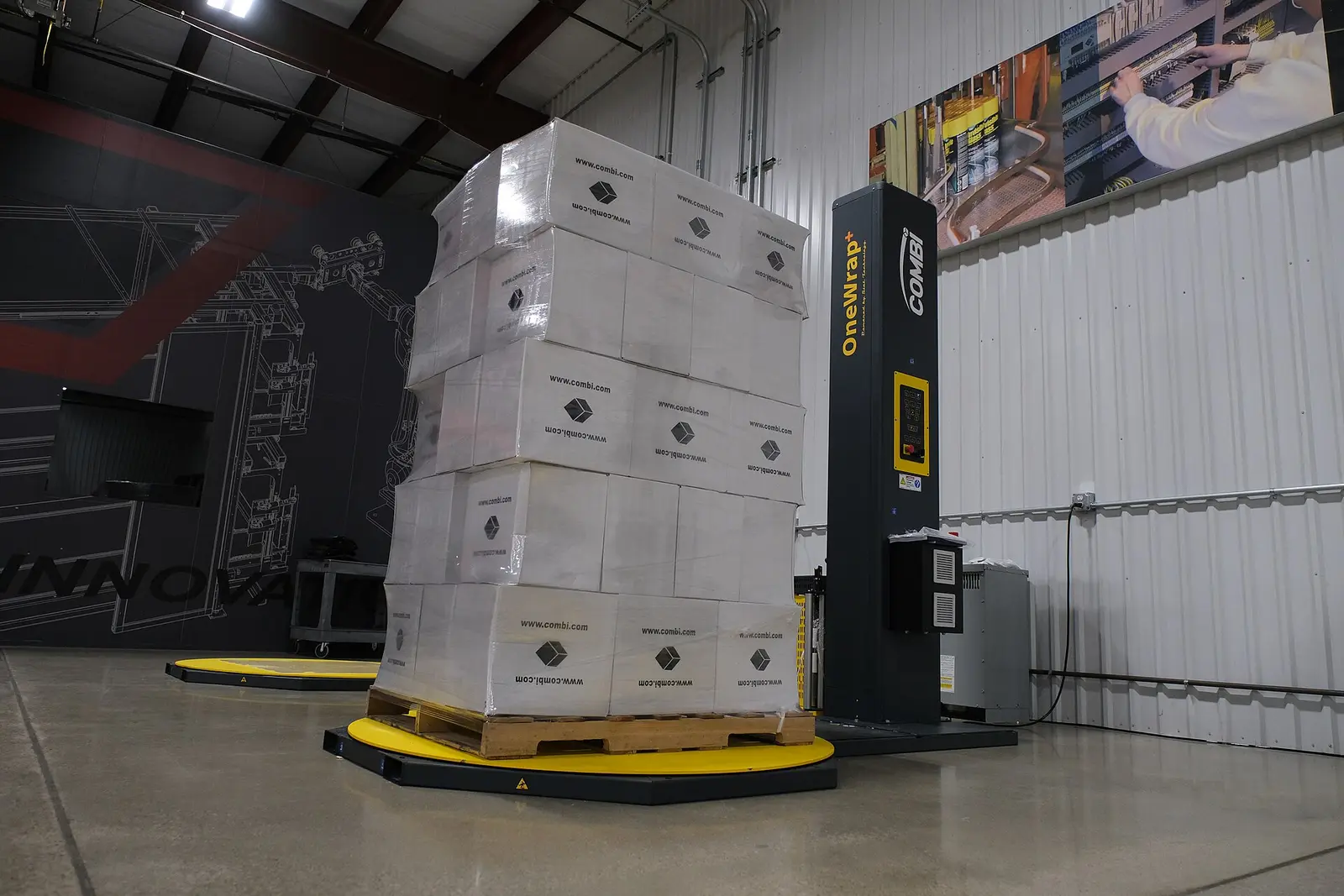 Turntable pallet stretch wrapper, with a film-wrapped pallet of cartons on the turntable beside the film carriage mast