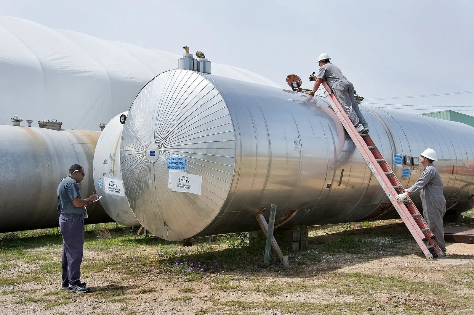 Steel chemical storage tanks, showing horizontal cylindrical steel vessels on saddle supports with top nozzles being inspected by workers