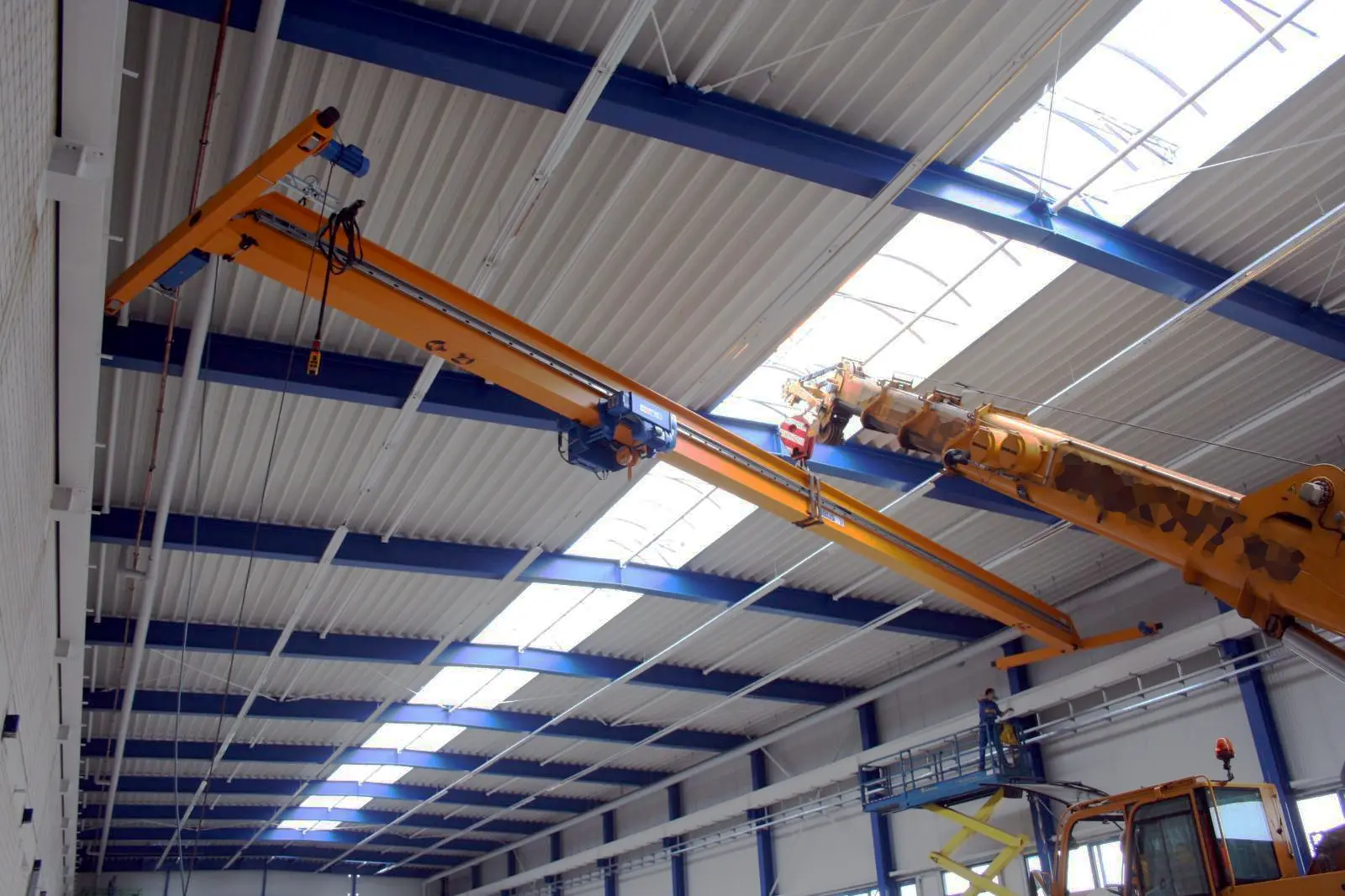 Orange single-girder overhead travelling crane with a blue electric hoist being lifted onto its runway beams under the roof of a new industrial workshop hall